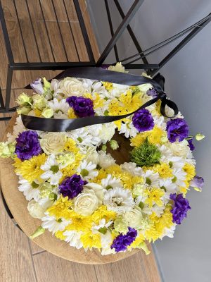 Coroană funerară rotundă cu crizanteme albe și galbene, trandafiri albi și eustoma mov, panglică neagră de satin